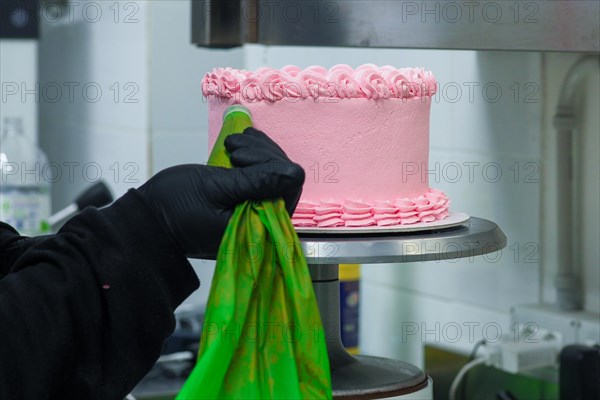 Pink cake being decorated with frosting from a green piping bag on a metal stand in a bakery by pastry chef usign black latex gloves