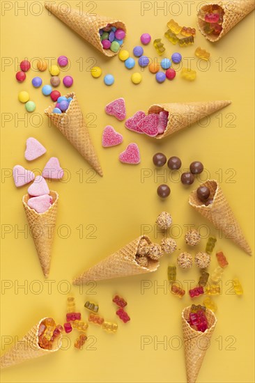 Colourful sweets, gummy bears and chocolate lentils fall out of ice cream cones on a yellow background