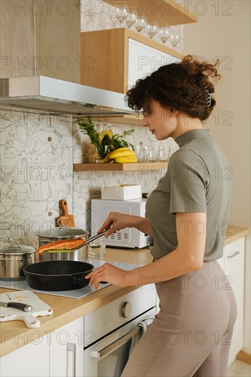 A woman takes a fried salmon steak out of a frying pan in her kitchen