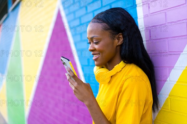 African woman smiling while using phone leaning on a multicolored wall in the city