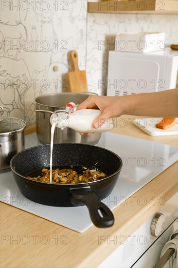 Unrecognizable woman pours cream into a frying pan with fried vegetables