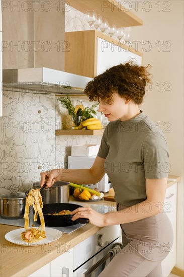 Young woman puts cooked pasta with mushroom sauce from a frying pan on a plate