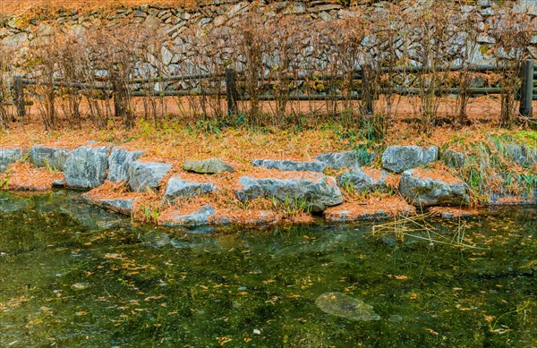 Boulders on shoreline of small frozen pond with wooden railing fence and stone wall in background in South Korea