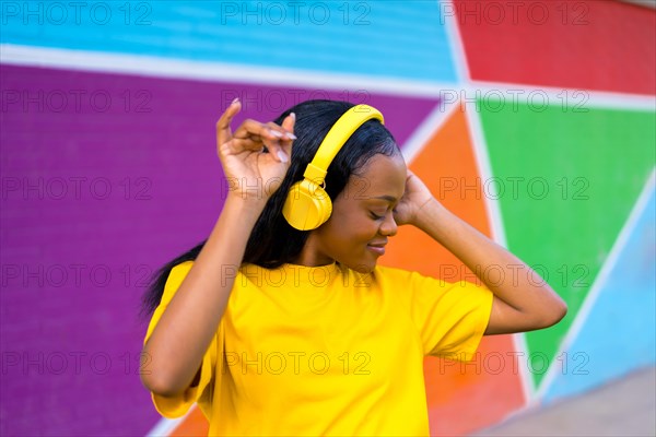 Portrait of a cool african young woman with yellow headphones and t-shirt dancing next to vivid colorful wall