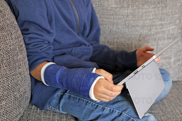 Boy with arm in plaster sits on the sofa with a tablet