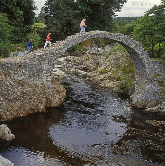 Three women walk across the packhorse bridge at Carrbridge in Scotland, Great Britain, Europe. Scanned 6x6 slide