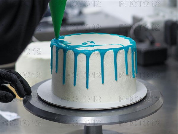 Applying final touches of blue icing to a freshly frosted white frosted cake in professional caterer kitchen