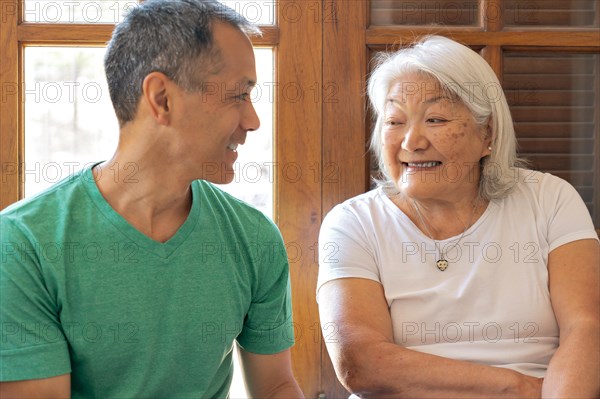 A grey-haired elderly mother of Japanese origin looks at her adult son smiling and sharing a happy moment at home. Mother's Day concept