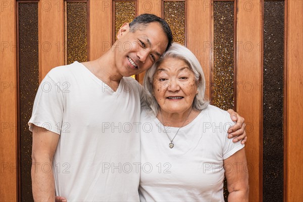 Happy older Asian mother and adult son hugging affectionately, looking at camera, smiling, enjoying family visit, free time