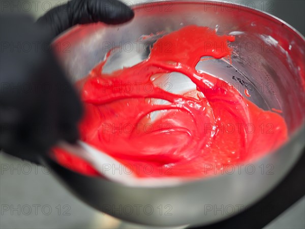 Pasty chef hands wearing black latex gloves stir on A shiny metal bowl full of bright red frosting being prepared for cake decorating