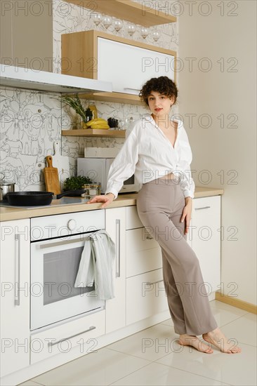 Barefoot young woman standing gracefully in a well-lit modern kitchen. She is wearing white shirt and grey pants complementing the sleek white cabinets and countertops