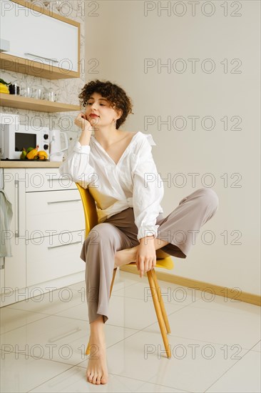 Young woman sits thoughtfully on a yellow chair in a well-lit, contemporary kitchen, embodying a moment of relaxation and contemplation