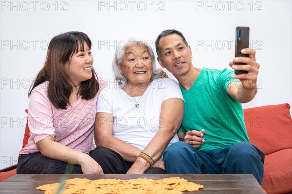Adult children and elderly white haired mother, ethnic Japanese family, take a selfie sitting on a sofa smiling and sharing a family moment