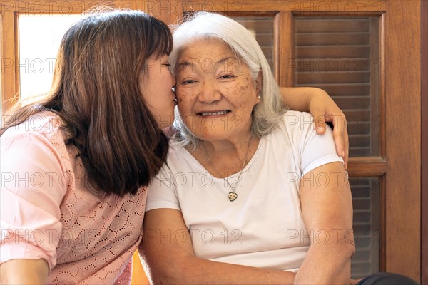 Affectionate Asian daughter kissing her mother at home. Affectionate adult daughter gently kisses the cheek of her Japanese mother with gray hair. Mothers Day