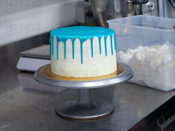 A white frosted cake with vibrant blue dripping icing on a cake stand ready for decoration