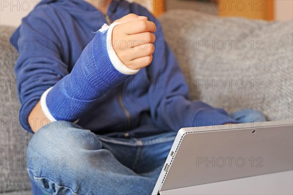 Boy with arm in plaster sits on the sofa with a tablet