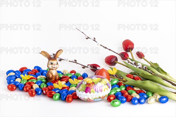 A collection of colourful chocolate eggs and an Easter bunny next to tulips and palm catkins, white background