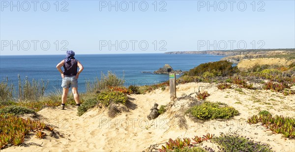 One woman walking on cliffs above the Atlantic Ocean on coastal long distance footpath trail, The Fisherman's Walk or Ruta Vicentina, near Zambujeira do Mar, Alentejo Littoral, Portugal, Southern Europe. To her side is the painted way-mark sign for the route, Europe