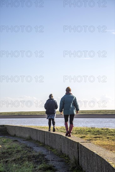 Mother and adult daughter walking on a low wall together by the River Ore, Suffolk, England, UK