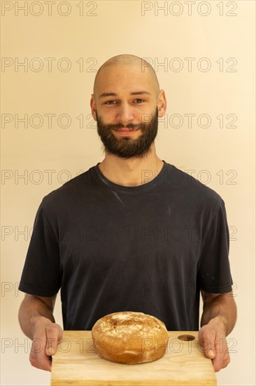 Head and shoulder portrait Caucasian white young man shaven head and beard holding fresh loaf of bread he has baked