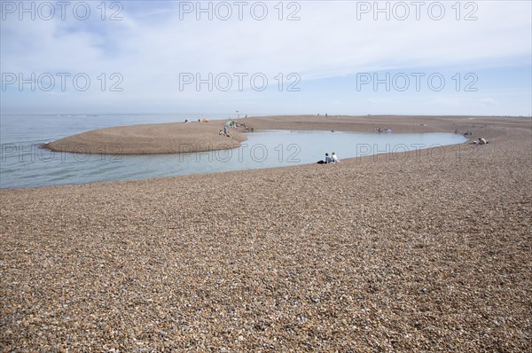 People enjoying a clam day sitting around a recently formed lagoon on the beach at Shingle Street, Suffolk, England, UK