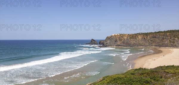 Atlantic Ocean waves breaking on rocky headland and bay with wide sandy beach, surfer and a few sunbathers, Praia de Odeceixe, Algarve, Portugal, Southern Europe, Europe