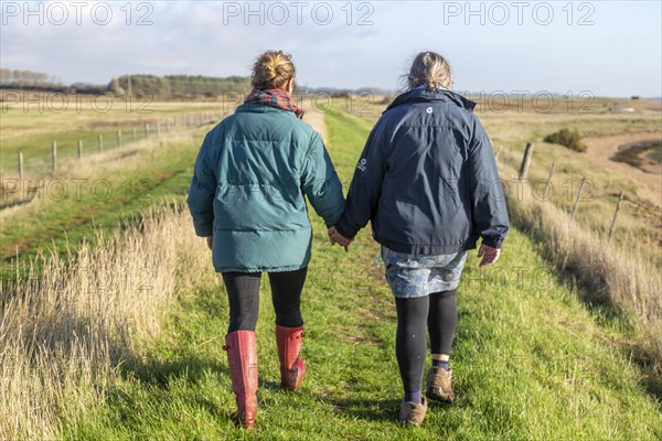 Mother and adult daughter walking together by the River Ore, Suffolk, England, UK from behind holding hands