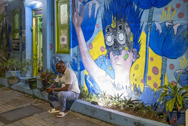 Man sits on a curb in front of illuminated street art in the streets of Fort Kochi, Cochin, Kerala, India, Asia