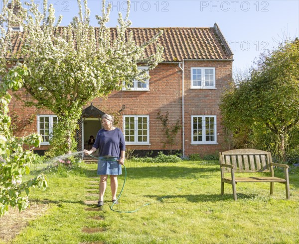 Woman watering garden with hosepipe in front of red brick historic detached house, Shottisham, Suffolk, England, UK