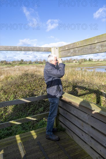 Male birdwatcher ornithologist using binoculars hide at RSPB Hollesley Marshes, Suffolk, England, UK
