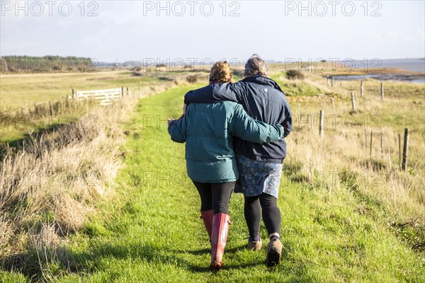 Mother and adult daughter walking together by the River Ore, Suffolk, England, UK from behind embracing