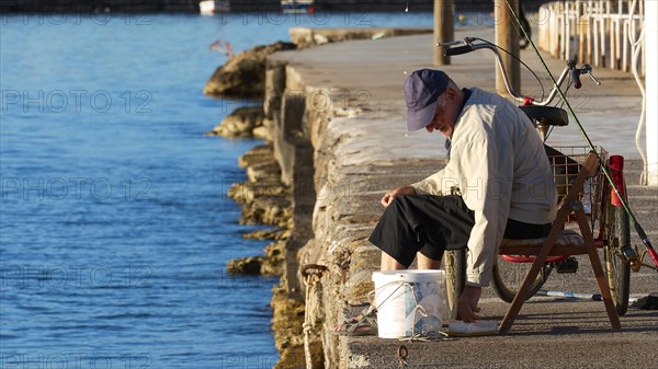 A man sitting on a folding chair fishing on the waterfront in the morning light, Gythio, Mani, Peloponnese, Greece, Europe