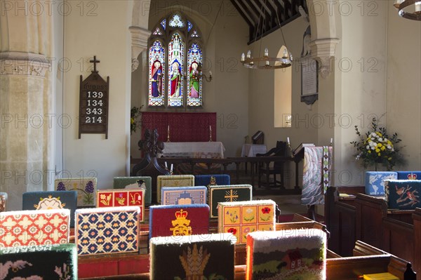 Knitted woollen pew kneelers rest on wooden pews, view towards altar and stained glass east window, interior of church at Stert, Wiltshire, England, UK