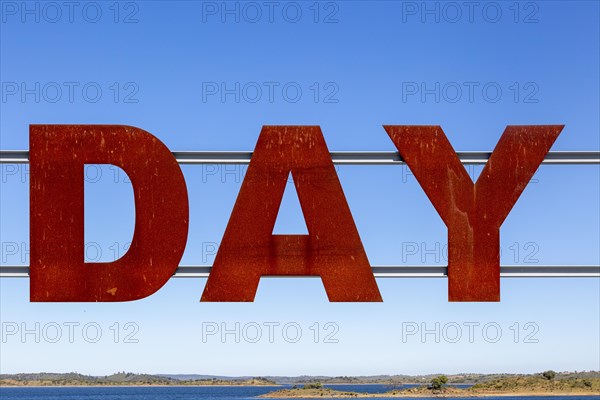 Large red rusty capital letters spell the word DAY against blue sky background, Alqueva dam, Moura, Portugal, Europe