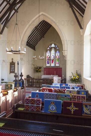 Knitted woollen pew kneelers rest on wooden pews, view towards altar and stained glass east window, interior of church at Stert, Wiltshire, England, UK