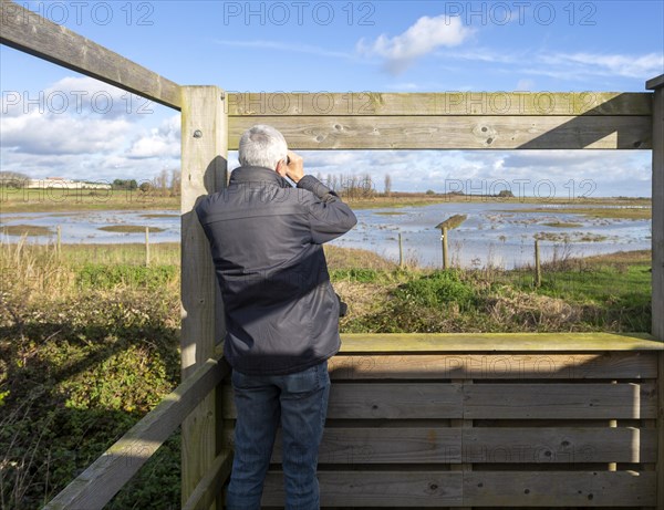 Male birdwatcher ornithologist using binoculars hide at RSPB Hollesley Marshes, Suffolk, England, UK