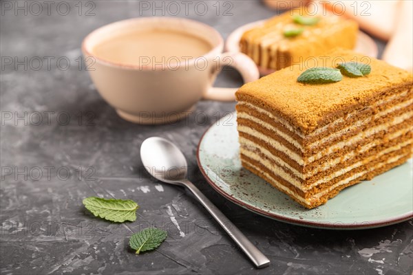 Homemade honey cake with milk cream and mint with cup of coffee on a black concrete background and orange textile. Side view, close up, selective focus