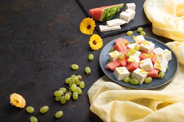 Vegetarian salad with watermelon, feta cheese, and grapes on blue ceramic plate on black concrete background and yellow linen textile. Side view, copy space