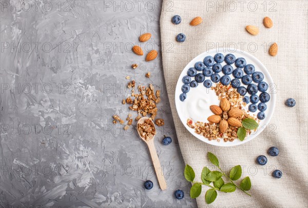Yoghurt with blueberry, granola and almond in white plate with wooden spoon on gray concrete background and linen textile. top view, flat lay, copy space