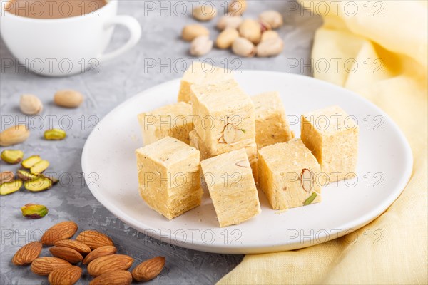 Traditional indian candy soan papdi in white plate with almond, pistache and a cup of coffee on a gray concrete background with yellow textile. side view, close up, selective focus