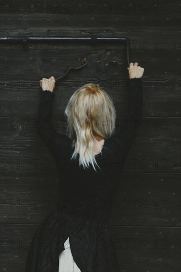 Back view of a blonde thin white woman dressed in black reaching up to a rusty antique metal lock on a wooden door with both hands