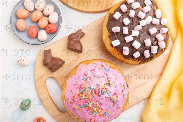 Homemade glazed and decorated easter pies with chocolate eggs and rabbits on a white concrete background and yellow textile. top view, flat lay, close up