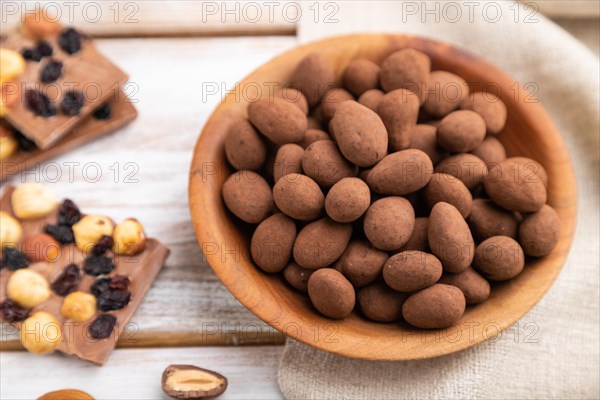 Almond in chocolate dragees in wooden plate and a cup of coffee on white wooden background and linen textile. Side view, close up, selective focus