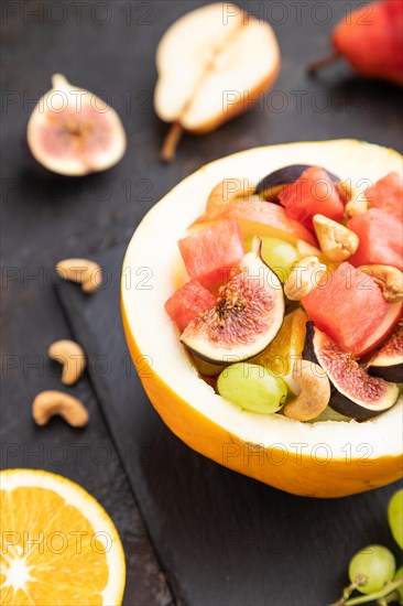 Vegetarian fruit salad of watermelon, grapes, figs, pear, orange, cashew on slate board on a black concrete background. Side view, close up, selective focus