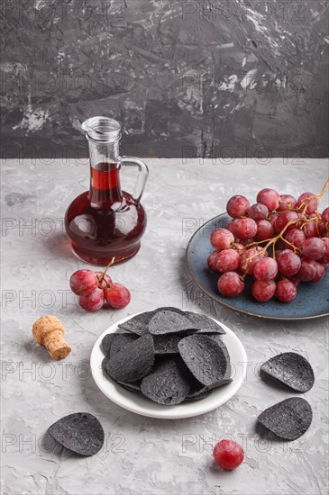 Black potato chips with charcoal, balsamic vinegar in glass, red grapes on a blue ceramic plate on a gray concrete background. side view, copy space, close up