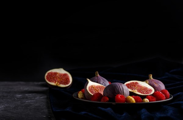 Fresh figs, strawberries and raspberries on blue ceramic plate on black concrete background and blue velvet textile. side view, close up, selective focus, low key