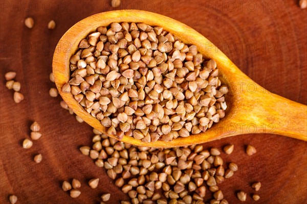 Pile of buckwheat in a wooden spoon on brown background. Closeup