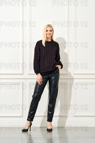 An elegant woman poses confidently against a white, panelled wall. She is dressed in a dark, patterned sweater and sleek black leather pants, complemented by classic black heels
