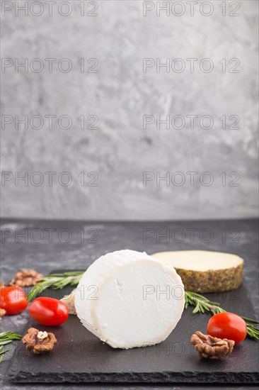 White goat cheese and various types of cheese with rosemary and tomatoes on black slate board on a black concrete background. Side view, copy space, selective focus