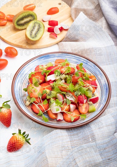 Vegetarian fruits and vegetables salad of strawberry, kiwi, tomatoes, microgreen sprouts on white concrete background and linen textile. Side view, close up